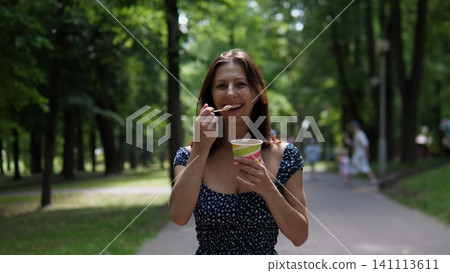 Smiling woman holding colorful ice cream cup and spoon on shaded park path, surrounded by lush green trees, enjoying relaxed summer leisure outdoors with soft natural daylight Smiling woman holding colorful ice cream cup and spoon on shaded park path, surrounded by lush green trees, enjoying relaxed summer leisure outdoors with soft natural daylight 141113611