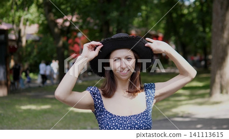 Smiling young woman adjusts black wide brim hat while standing outdoors in sunny park, wearing blue floral dress, with soft daylight, blurred trees, green lawn, relaxed summer mood 141113615