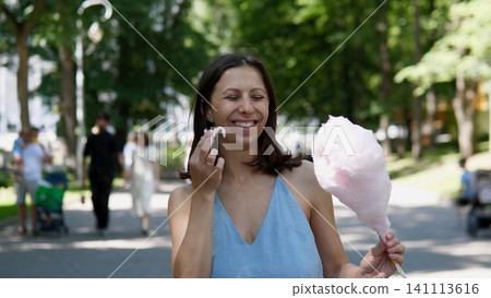 Young woman tasting pink cotton candy in bright green park, surrounded by soft focus trees and visitors, showing cheerful summer leisure, outdoor recreation and playful fair 141113616