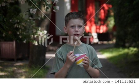Young boy standing on shaded garden path, holding striped cup of ice cream and spoon, with soft blurred flowers, greenery, red building and gentle summer daylight behind him Young boy standing on shaded garden path, holding striped cup of ice cream and spoon, with soft blurred flowers, greenery, red building and gentle summer daylight behind him 141113621