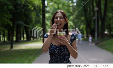 Cheerful young woman tastes ice cream from paper cup beside park path surrounded by green trees, sunlit summer scenery, soft bokeh, and relaxed outdoor mood, with woman, smiling Cheerful young woman tastes ice cream from paper cup beside park path surrounded by green trees, sunlit summer scenery, soft bokeh, and relaxed outdoor mood, with woman, smiling 141113630