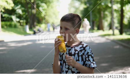 Young boy in patterned shirt eating fresh corn on cob beside tree lined park path, surrounded by bright greenery, soft sunlight, blurred walkers in background, relaxed summer Young boy in patterned shirt eating fresh corn on cob beside tree lined park path, surrounded by bright greenery, soft sunlight, blurred walkers in background, relaxed summer 141113636