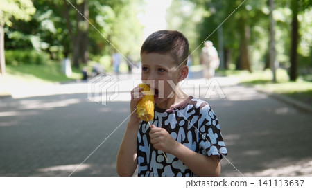 Child enjoys fresh corn on cob in sunny city park, holding ear with both hands beside tree-lined walkway, soft bokeh background and warm daylight create relaxed summer mood Child enjoys fresh corn on cob in sunny city park, holding ear with both hands beside tree-lined walkway, soft bokeh background and warm daylight create relaxed summer mood 141113637