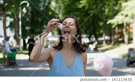 Smiling woman in blue dress tasting cotton candy in leafy park, standing on sunlit path with soft bokeh background, carefree summer moment, casual outdoor leisure and fun Smiling woman in blue dress tasting cotton candy in leafy park, standing on sunlit path with soft bokeh background, carefree summer moment, casual outdoor leisure and fun 141113638