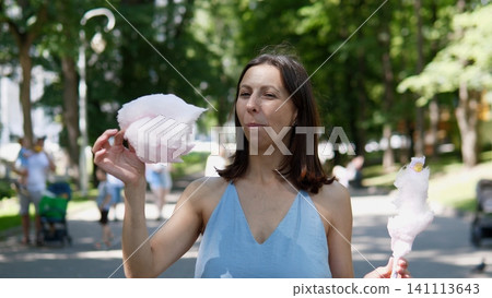 Young woman in blue sleeveless top holds fluffy cotton candy in sunny park, with trees, pathway, and blurred visitors behind her, creating relaxed summer outdoor leisure scene Young woman in blue sleeveless top holds fluffy cotton candy in sunny park, with trees, pathway, and blurred visitors behind her, creating relaxed summer outdoor leisure scene 141113643