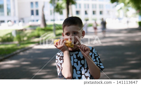 Child enjoys grilled corn outdoors on sunny summer walkway, holding cob with both hands near mouth, while soft focus background shows trees, building, and passing people Child enjoys grilled corn outdoors on sunny summer walkway, holding cob with both hands near mouth, while soft focus background shows trees, building, and passing people 141113647