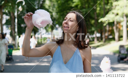 Smiling woman in blue sleeveless top holds fluffy pink cotton candy beside face on bright park path with sunlit trees, blurred visitors, and soft natural outdoor light Smiling woman in blue sleeveless top holds fluffy pink cotton candy beside face on bright park path with sunlit trees, blurred visitors, and soft natural outdoor light 141113722