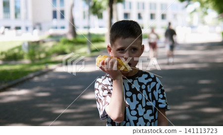 Young boy enjoys roasted corn on cob while standing on shaded park path with sunlit trees, blurred buildings, and distant people creating calm summer outdoor scene Young boy enjoys roasted corn on cob while standing on shaded park path with sunlit trees, blurred buildings, and distant people creating calm summer outdoor scene 141113723