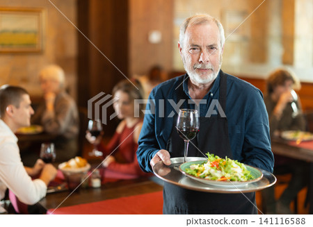 Elderly male waiter with order on tray in restaurant Elderly male waiter with order on tray in restaurant 141116588