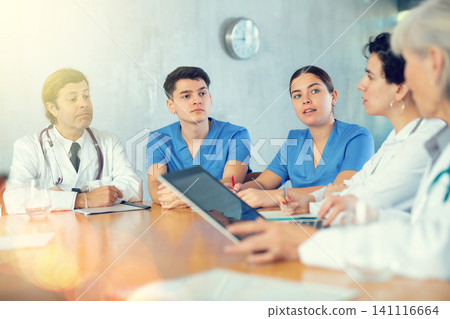 Doctors and nurses of different ages discussing topic of day while sitting at table with laptop and notes Doctors and nurses of different ages discussing topic of day while sitting at table with laptop and notes 141116664