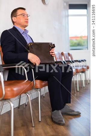 Serious-minded male candidate siting on chair and waiting for job interview in hallway Serious-minded male candidate siting on chair and waiting for job interview in hallway 141116695