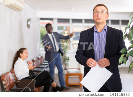 Upset middle-aged man standing in waiting room with his back to person who is shouting at him Upset middle-aged man standing in waiting room with his back to person who is shouting at him 141116883