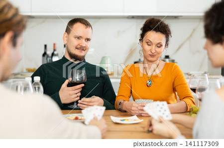 Friendly company of men and women sitting at kitchen table and enjoying the poker game. Friends play a card game at home. Evening entertainment concept Friendly company of men and women sitting at kitchen table and enjoying the poker game. Friends play a card game at home. Evening entertainment concept 141117309