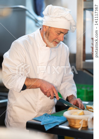 Eldery male chef slicing bread in kitchen Eldery male chef slicing bread in kitchen 141117367
