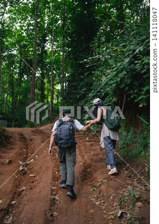 Two hikers with backpacks holding hands walking up dirt path in dense green forest during outdoor adventure Two hikers with backpacks holding hands walking up dirt path in dense green forest during outdoor adventure 141118047