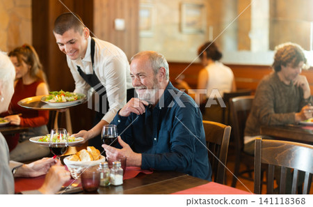 Young waiter bringing food to elderly couple in restaurant Young waiter bringing food to elderly couple in restaurant 141118368