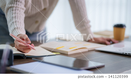 Woman hand writing notes in notebook at wooden desk with laptop and coffee in bright home office Woman hand writing notes in notebook at wooden desk with laptop and coffee in bright home office 141120399