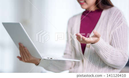 Young Asian woman presenting online education tutorial holding laptop and gesturing in bright home office with copy space 141120410