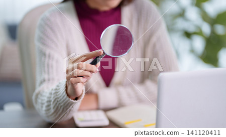Adult woman holding magnifying glass while studying or researching with laptop and notebook in bright home office Adult woman holding magnifying glass while studying or researching with laptop and notebook in bright home office 141120411