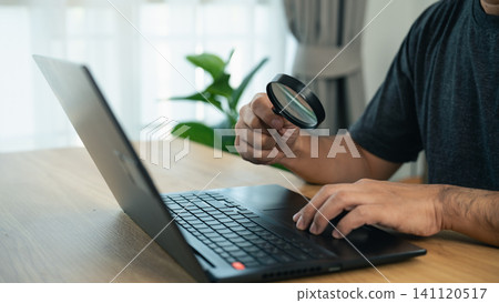Person examining laptop screen closely with magnifying glass sitting at wooden table in bright room with natural light Person examining laptop screen closely with magnifying glass sitting at wooden table in bright room with natural light 141120517
