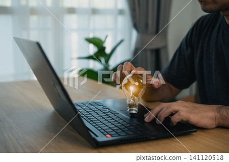 Man holding illuminated light bulb while using laptop on wooden table indoors next to window with natural light and plant in background 141120518