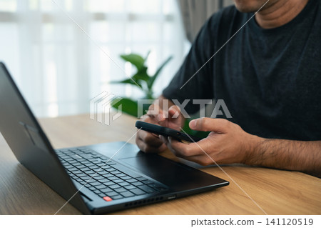 Man using smartphone and laptop at wooden table in bright modern room with natural light and casual clothing focusing on device interaction Man using smartphone and laptop at wooden table in bright modern room with natural light and casual clothing focusing on device interaction 141120519