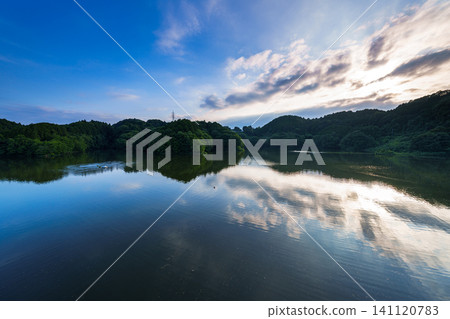 Nunome Dam in eastern Nara City: The sunset sky is reflected on the calm surface of the lake, and the setting sun is hidden by clouds drifting in from the southwest. 141120783