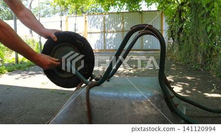 A man repairs an overturned construction wheelbarrow in a yard against a fence. His hands unscrew an old, worn wheel with a blue disc from the metal axle of the frame, removing the mounting bolt. A man repairs an overturned construction wheelbarrow in a yard against a fence. His hands unscrew an old, worn wheel with a blue disc from the metal axle of the frame, removing the mounting bolt. 141120994
