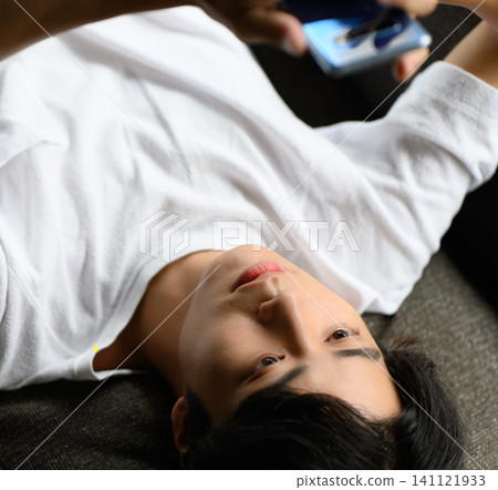A young man is lying on a sofa, staring at his smartphone. A young man is lying on a sofa, staring at his smartphone. 141121933