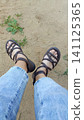 First-person top-down view of feet wearing strappy brown sandals and blue jeans, standing on sandy ground with green weeds. 141125365
