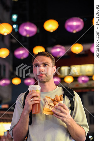 Handsome 30 years old Russian man tourist holding traditional bubble waffle and iced lemon tea at illuminated Temple Street Night Market in Hong Kong 141126692