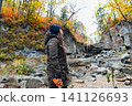 Russian solo traveler woman standing sideways with smartphone near flowing waterfall in autumn forest at Kravtsovskie waterfalls travel nature concept 141126693