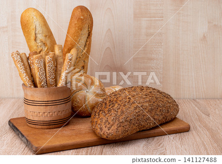 Crispy bread sticks with sesame seeds and bran bread on a wooden board. French baguettes. Different breeds on wooden background. Crispy bread sticks with sesame seeds and bran bread on a wooden board. French baguettes. Different breeds on wooden background. 141127488