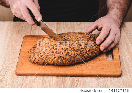 A man with a knife cuts bread on a wooden board. 141127566