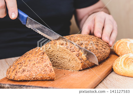 A man with a knife cuts bread on a wooden board. French baguettes. Different breeds on wooden background. A man with a knife cuts bread on a wooden board. French baguettes. Different breeds on wooden background. 141127583