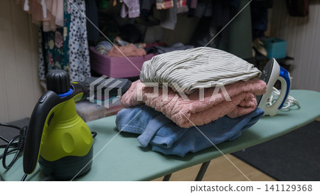 Tidy clothing stack on ironing board, iron and handheld steamer set housework concept. Steaming equipment. Folded clothes and tools. Meticulously arranged laundry space nook. Casual domestic routine 141129368