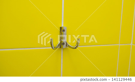 Silver metal cloth on tiled yellow wall. Strong Steel Wall Hanger. Chrome Hanger hook mounted on tile background. Mock-up. Iron clothes hook is mounted on a light wall. Ideal for hanging clothes. Silver metal cloth on tiled yellow wall. Strong Steel Wall Hanger. Chrome Hanger hook mounted on tile background. Mock-up. Iron clothes hook is mounted on a light wall. Ideal for hanging clothes. 141129446