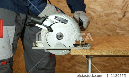 Builder saws a board with a circular saw cutting a wooden plank in a joinery. Carpenter uses portable circular saw tool to cut wood at wooden table in workshop. Worker sawing pine board. Real image 141129555