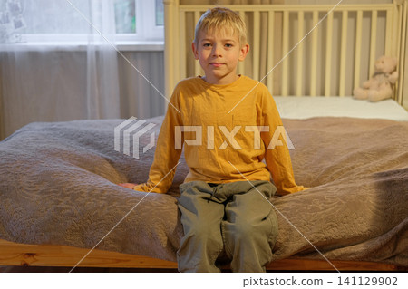 Young kid at home. Tranquil boy sitting comfortably. Content young boy relaxing peacefully with teddy bear toy. Calm and happy child. Cozy bedroom portrait Caucasian blond hair boy, warm atmosphere 141129902