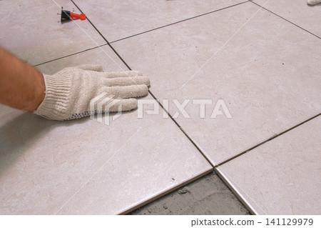 Worker Hand Laying Ceramic Tiles on the Floor. Repairman Placing White Gray Square Tiles, Applying Adhesive. Builder Installing Tile Using Leveler, Tile Leveling Clips. Tiling. Finishing Repair Work. Worker Hand Laying Ceramic Tiles on the Floor. Repairman Placing White Gray Square Tiles, Applying Adhesive. Builder Installing Tile Using Leveler, Tile Leveling Clips. Tiling. Finishing Repair Work. 141129979