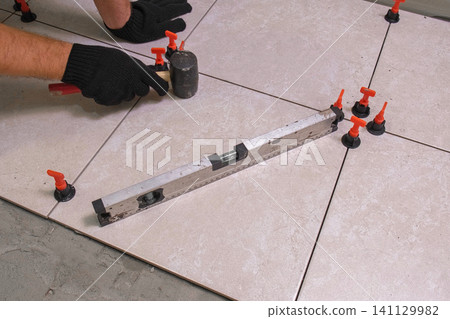 Worker Hand Laying Ceramic Tiles on the Floor. Repairman Placing White Gray Square Tiles, Applying Adhesive. Builder Installing Tile Using Leveler, Tile Leveling Clips. Tiling. Finishing Repair Work. Worker Hand Laying Ceramic Tiles on the Floor. Repairman Placing White Gray Square Tiles, Applying Adhesive. Builder Installing Tile Using Leveler, Tile Leveling Clips. Tiling. Finishing Repair Work. 141129982