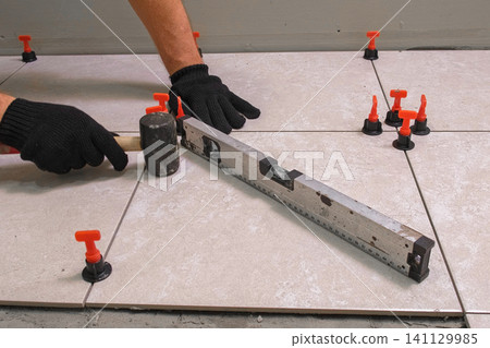 Worker Hand Laying Ceramic Tiles on the Floor. Repairman Placing White Gray Square Tiles, Applying Adhesive. Builder Installing Tile Using Leveler, Tile Leveling Clips. Tiling. Finishing Repair Work. Worker Hand Laying Ceramic Tiles on the Floor. Repairman Placing White Gray Square Tiles, Applying Adhesive. Builder Installing Tile Using Leveler, Tile Leveling Clips. Tiling. Finishing Repair Work. 141129985