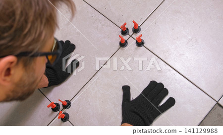 Worker Hand Laying Ceramic Tiles on the Floor. Repairman Placing White Gray Square Tiles, Applying Adhesive. Builder Installing Tile Using Leveler, Tile Leveling Clips. Tiling. Finishing Repair Work. Worker Hand Laying Ceramic Tiles on the Floor. Repairman Placing White Gray Square Tiles, Applying Adhesive. Builder Installing Tile Using Leveler, Tile Leveling Clips. Tiling. Finishing Repair Work. 141129988