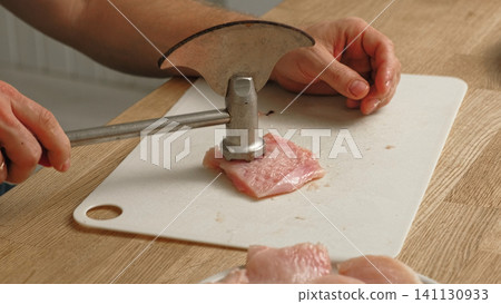 Person using meat tenderizer on raw chicken. Chef preparing pork loin slice with meat mallet on cutting board. Man tenderizing fillet. Cooking cutlets pounding poultry breast close-up. Culinary dish. Person using meat tenderizer on raw chicken. Chef preparing pork loin slice with meat mallet on cutting board. Man tenderizing fillet. Cooking cutlets pounding poultry breast close-up. Culinary dish. 141130933