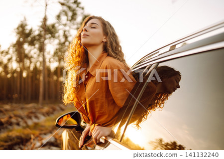 A young woman feels freedom leaning out of a car window in a sunny forest. Travel concept. A young woman feels freedom leaning out of a car window in a sunny forest. Travel concept. 141132240
