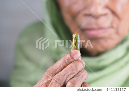 Elderly woman holding a small fish oil capsule with care Elderly woman holding a small fish oil capsule with care 141133157