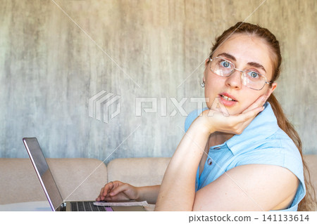 Young woman in glasses working on a laptop at home, looking thoughtful and slightly distracted Young woman in glasses working on a laptop at home, looking thoughtful and slightly distracted 141133614