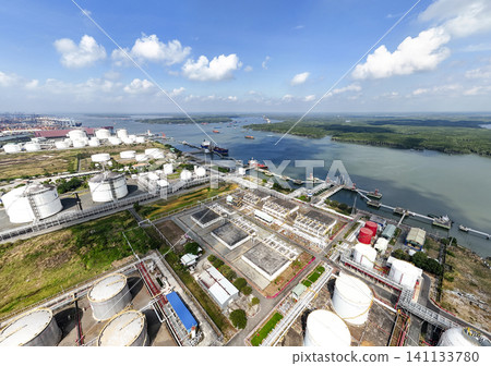 High-angle aerial drone view of a massive coastal industrial complex featuring oil and gas storage tanks, refinery pipelines, and cargo ports under a bright blue sky. 141133780