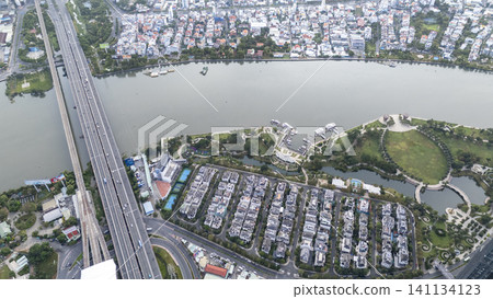 High-angle aerial panorama of a modern luxury residential district, featuring a major highway bridge and a lush riverside park along the city river. High-angle aerial panorama of a modern luxury residential district, featuring a major highway bridge and a lush riverside park along the city river. 141134123