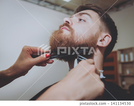 Master cuts hair and beard of men, hairdresser makes hairstyle for a young man. Hipsters Master cuts hair and beard of men, hairdresser makes hairstyle for a young man. Hipsters 141134530